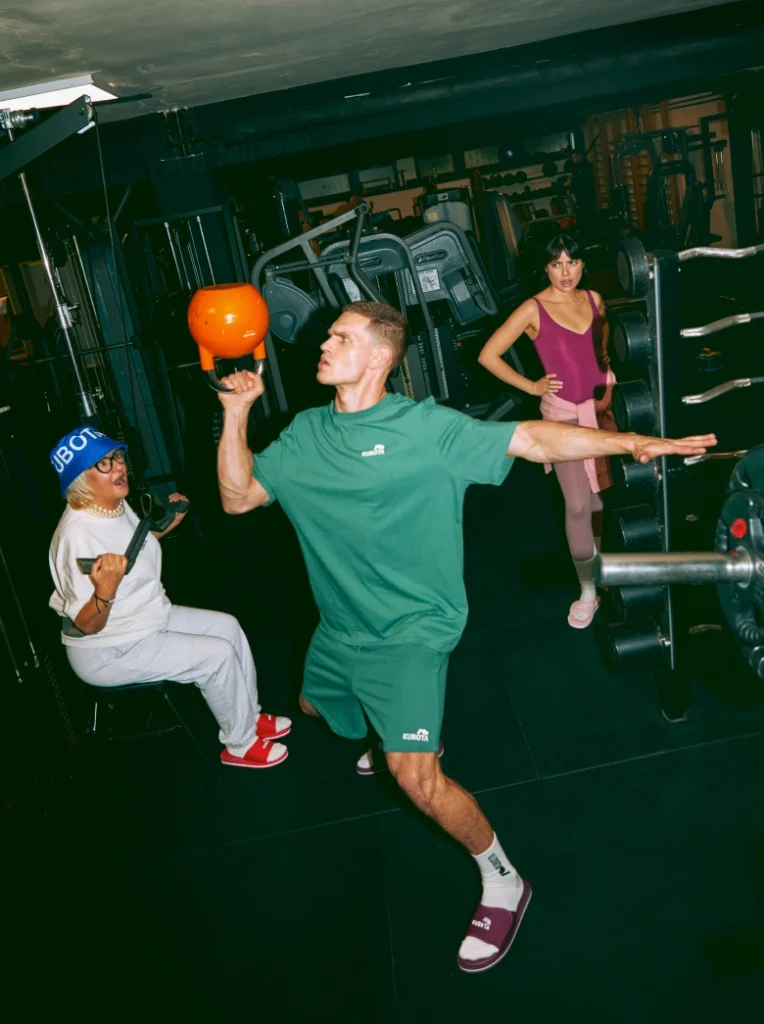 Model prezentuje stylizację lookbookową Kubota na siłowni, idealną na codzienną rozgrzewkę. Zestaw składa się z zielonego T-shirtu sportowego Move, zielonych szortów sportowych Move oraz burgundowych klapków Rzep.