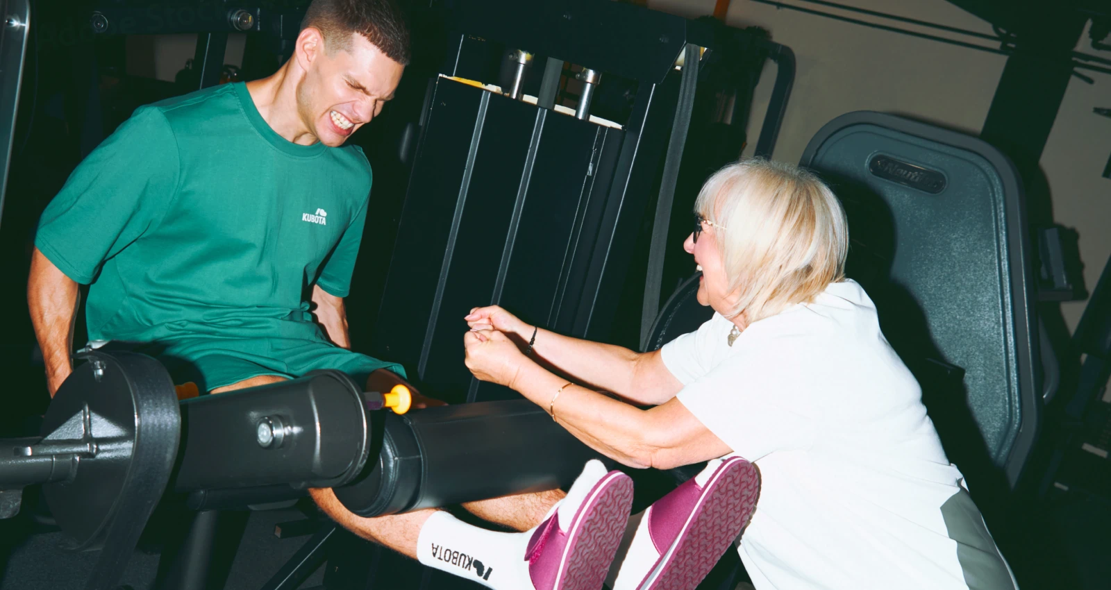 Modele prezentują stylizację lookbookową Kubota w trakcie codziennej rozgrzewki na siłowni. Mężczyzna nosi zielony T-shirt i szorty sportowe Move, a kobieta wspiera go w burgundowych klapkach Rzep.