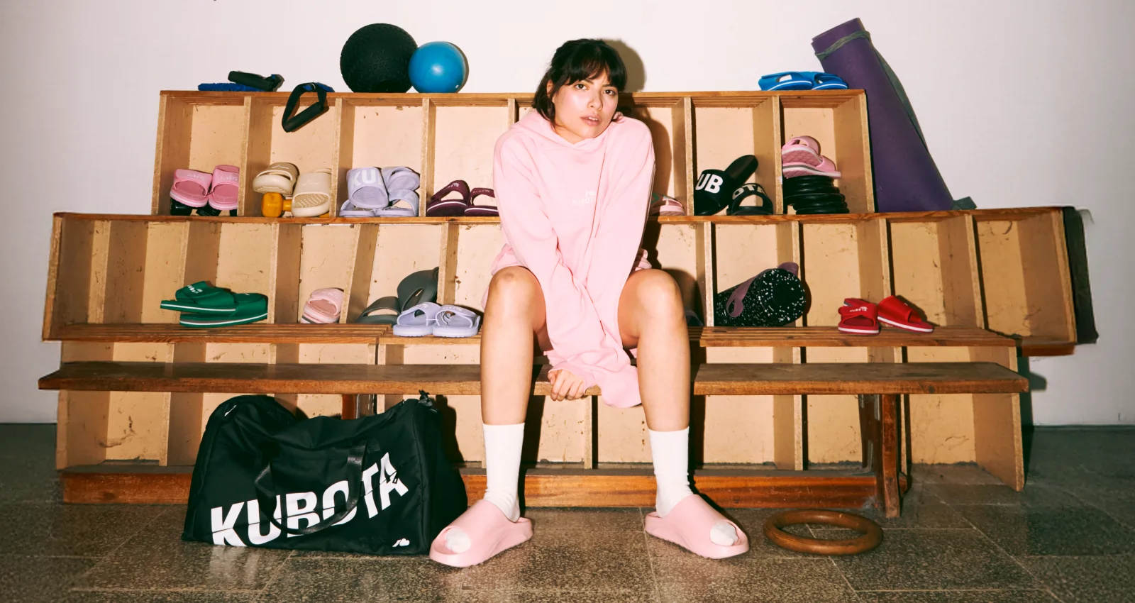 Modelka prezentuje stylizację lookbookową Kubota na siłowni, idealną na codzienną rozgrzewkę. Zestaw składa się z różowej bluzy z kapturem Statement, różowych szortów bawełnianych Statement i różowych klapków Flow.