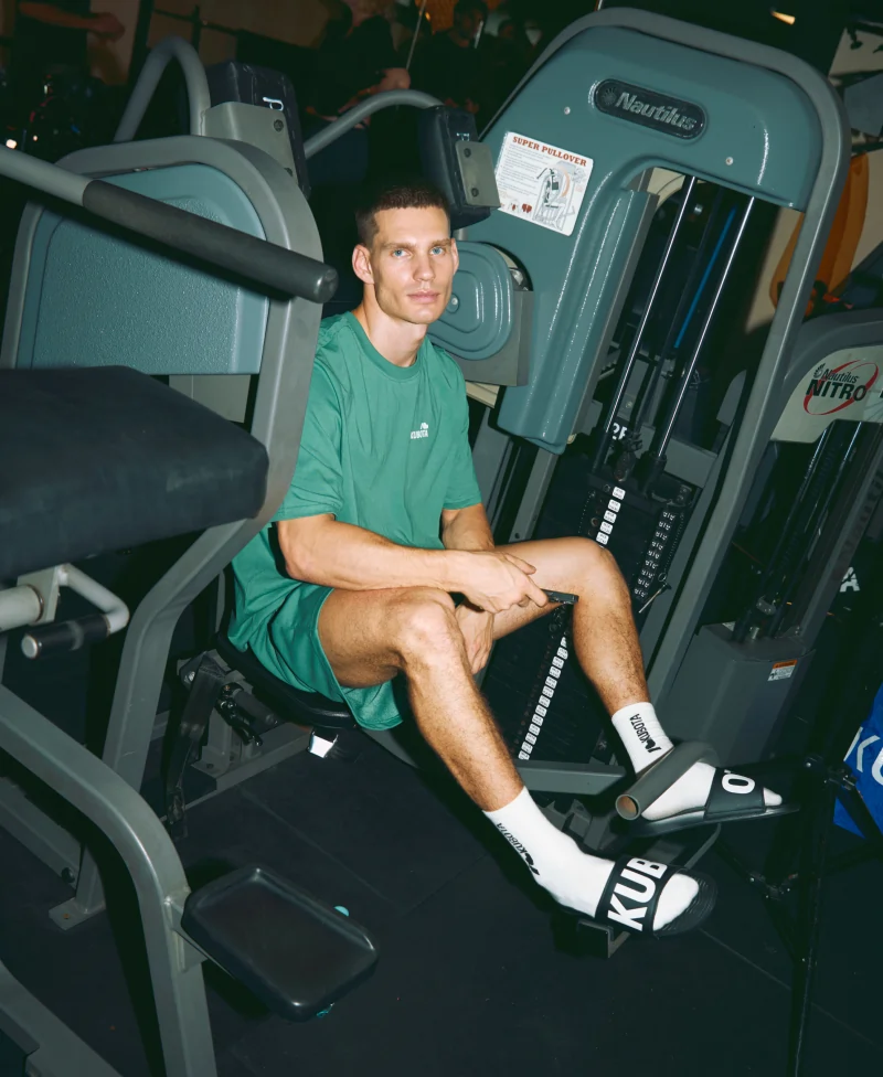 Model prezentuje stylizację lookbookową Kubota na siłowni, idealną na codzienną rozgrzewkę. Zestaw składa się z zielonego T-shirtu sportowego Move, zielonych szortów sportowych Move i czarnych klapków Pro 2.0.