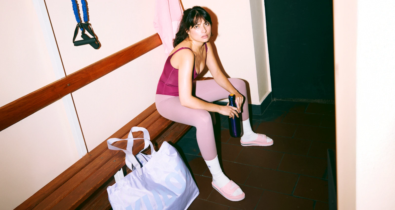 Modelka prezentuje stylizację lookbookową Kubota w szatni, idealną na codzienną rozgrzewkę. Zestaw składa się z pastelowo różowych klapków Rzep, kremowych skarpet Cloud oraz fioletowej torby Shopper Festiwal.