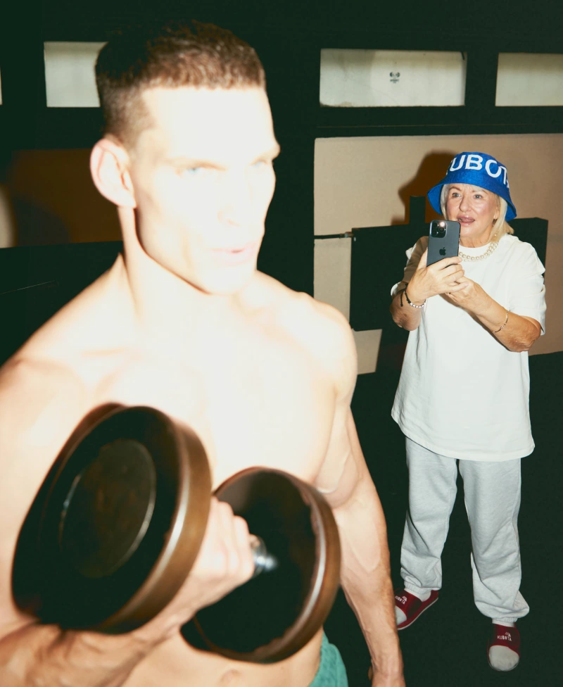 Modele prezentują scenę lookbookową Kubota na siłowni w trakcie codziennej rozgrzewki. Kobieta w kobaltowym kapeluszu Bucket Hat Festiwal, T-shircie Statement ecru i czerwonych klapkach Rzep robi zdjęcie mężczyźnie ćwiczącemu z hantlami.