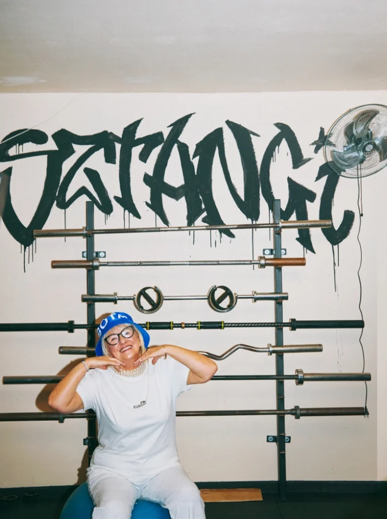 Modelka prezentuje stylizację lookbookową Kubota na siłowni, siedząc na piłce gimnastycznej. Zestaw na codzienną rozgrzewkę składa się z T-shirtu bawełnianego Statement ecru i kobaltowego kapelusza Bucket Hat Festiwal.