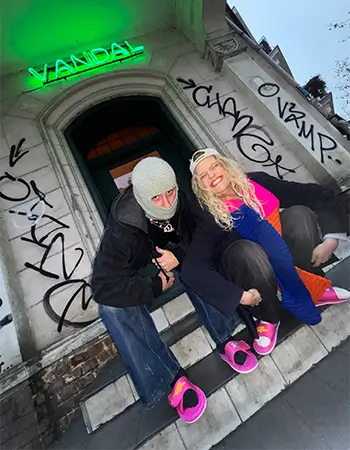 Zdjęcie @easytiger.agency w streetwearowym wydaniu. Dwie osoby w różowych klapkach Kubota Rzep na tle neonu VANDAL i graffiti.