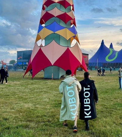 Zdjęcie @ka_lampone na festiwalu muzycznym. Dwie osoby w parkach przeciwdeszczowych z dużym logo Kubota na plecach na tle kolorowych instalacji.