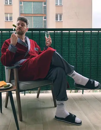 Zdjęcie @make-life-harder w czerwonym szlafroku, czarnych klapkach Kubota Rzep i białych skarpetkach, trzymającego ogórka kiszonego i kieliszek na balkonie.