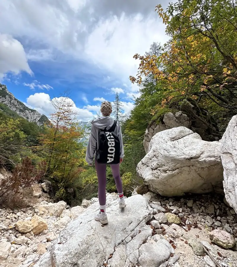 Zdjęcie @niejedzznami na górskim szlaku. Kobieta w szarej bluzie i czarnym plecaku z dużym, białym logo Kubota spogląda na góry.