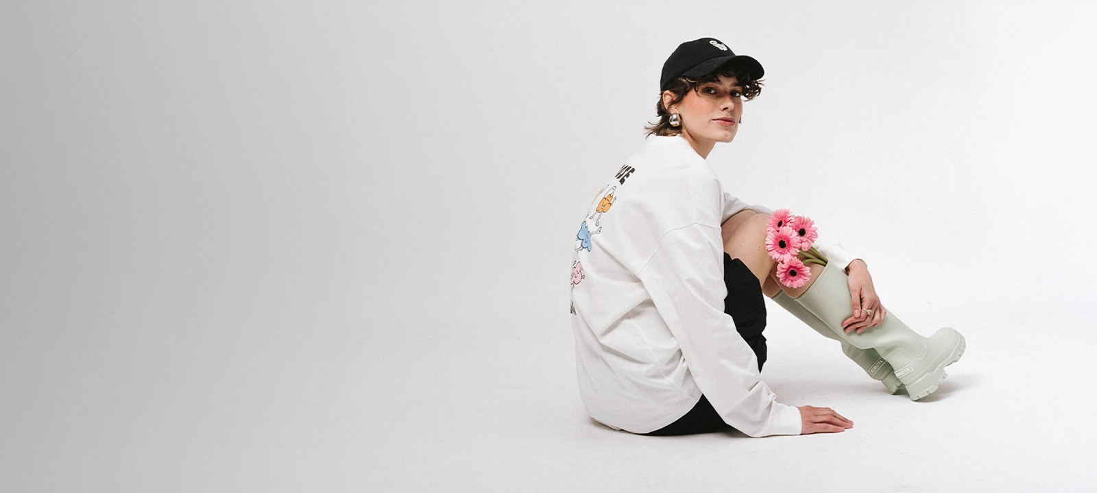 Baner kategorii Nowości. Modelka w białym longsleeve z grafiką, wysokich kaloszach w kolorze matchy i czarnej czapce od Kuboty z nowej kolekcji.