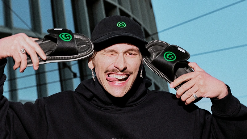 Baner kategorii NEXT FEST Merch. Model trzymający klapki Rzep NEXT FEST i noszący również czapkę z tej kolaboracji.