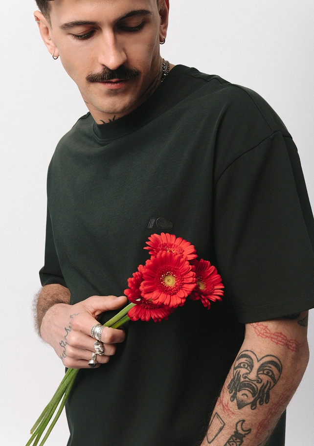 Zdjęcie lookbookowe z kolekcji SS26 prezentujące męski, ciemnozielony t-shirt Easy. Model z tatuażami trzyma w dłoni bukiet czerwonych kwiatów na jasnym tle.