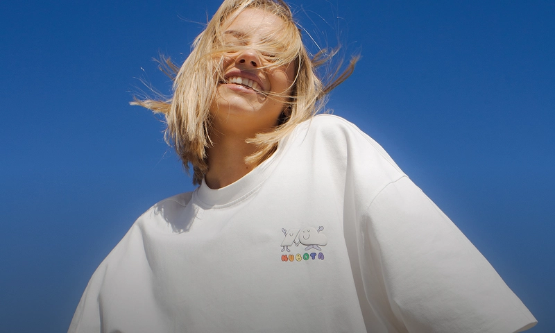 Baner kategorii bestsellery. Uśmiechnięta kobieta w białym t-shircie z logo na tle błękitnego nieba.