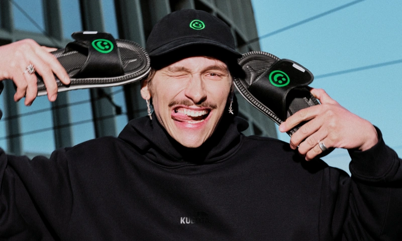 Baner podstrony Kubota na NEXT FEST. Model trzymający klapki Rzep NEXT FEST i noszący również czapkę z tej kolaboracji.