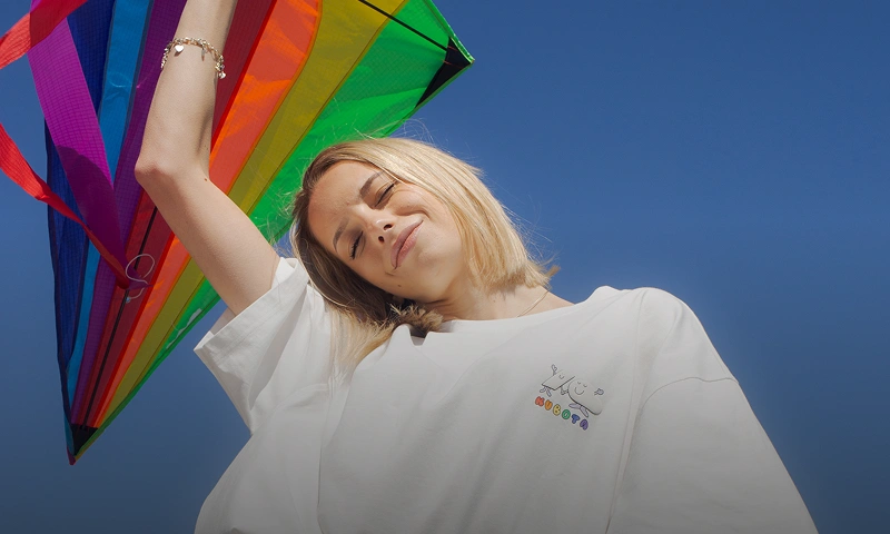 Baner kategorii Pride Month. Kobieta w białym t-shircie Comfy Club Pride trzyma tęczowy latawiec na tle błękitnego nieba.