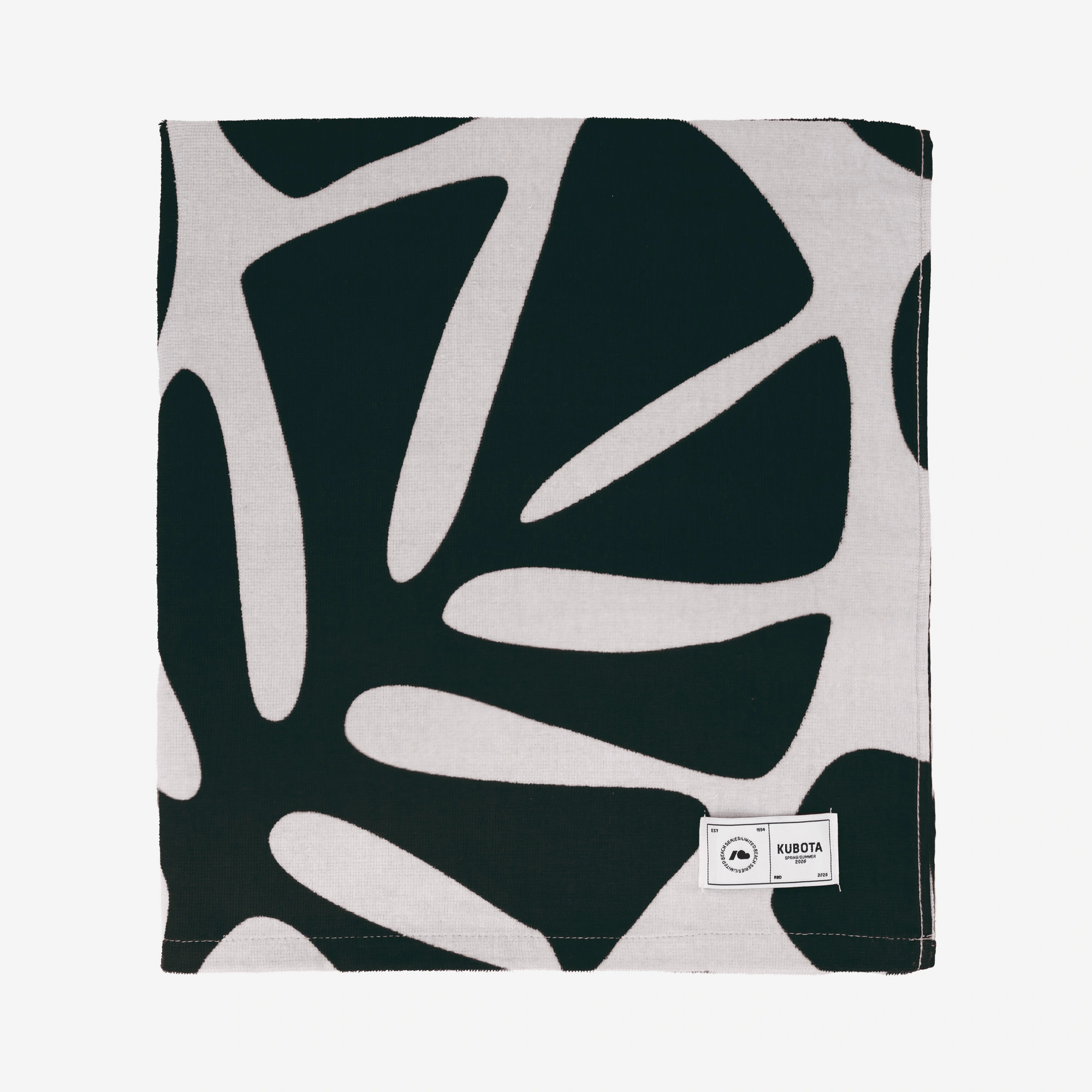 Zdjęcie katalogowe prezentujące ciemnozielono-różowy ręcznik plażowy z limitowanej kolekcji Kubota w Rossmannie. Artykuł zaprezentowany na białym tle wyróżnia się dużym motywem roślinnym oraz prostokątną, białą metką z czarnym logo marki.