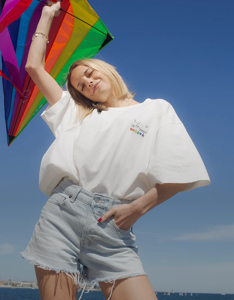 Grafika wizerunkowa na podstronie o zaangażowaniu społecznym. Uśmiechnięta kobieta w białym t-shircie Comfy Club Pride z kolorowym, tęczowym logo trzyma tęczowy latawiec na tle czystego, błękitnego nieba.