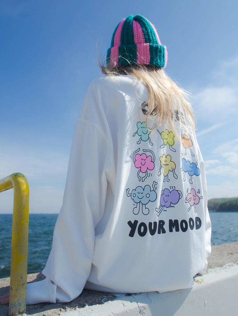 Zdjęcie lookbookowe nad morzem. Widok z tyłu na osobę w białym longsleeve oversize Comfy Club Moods oraz w pasiastej, różowo-zielonej czapce.
