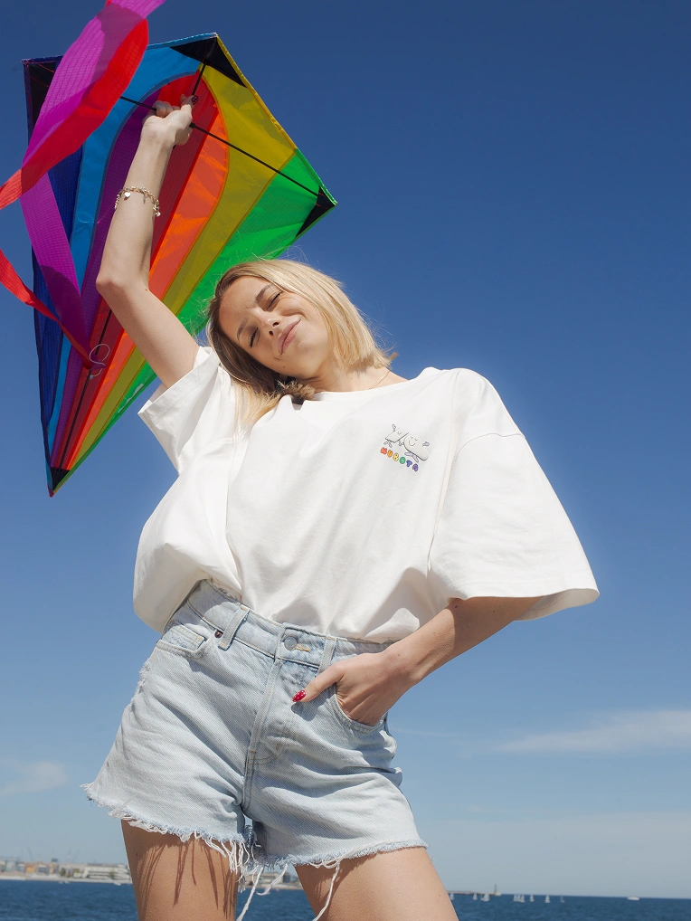 Zdjęcie lookbookowe uśmiechniętej kobiety w białym t-shircie Comfy Club Pride z kolorowym, tęczowym logo trzymającej tęczowy latawiec na tle czystego, błękitnego nieba.