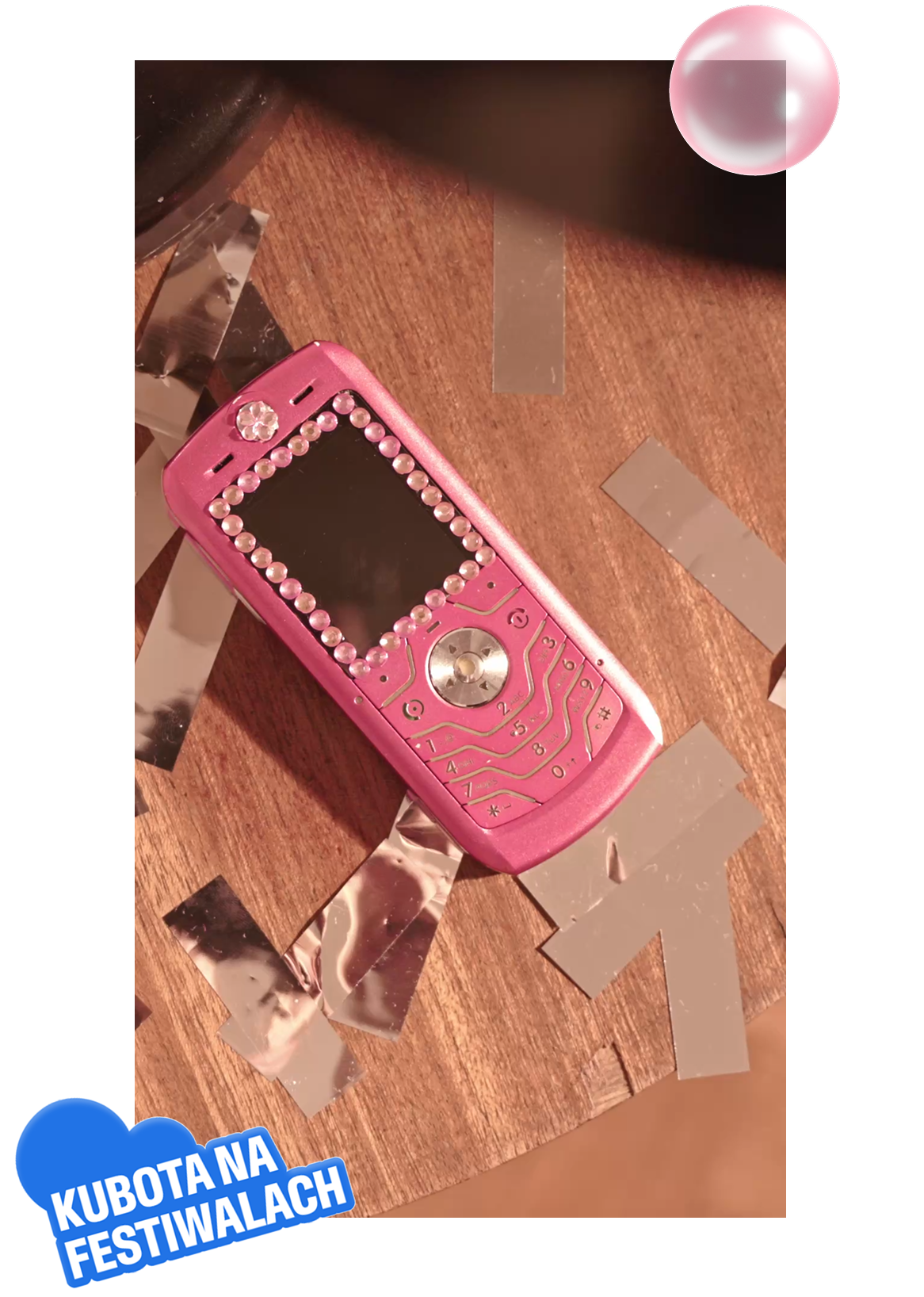 Różowy klasyczny telefon komórkowy ozdobiony błyszczącymi cyrkoniami leży na drewnianym blacie w otoczeniu srebrnego confetti. W rogu kadru widnieje niebieski akcent graficzny z napisem Kubota Na Festiwalach. Dedykowany rekwizyt w klimacie retro buduje nostalgiczny nastroj wokół projektu Kubota na BitterSweet, stanowiąc element unikalnej oprawy wizualnej letniego wydarzenia.