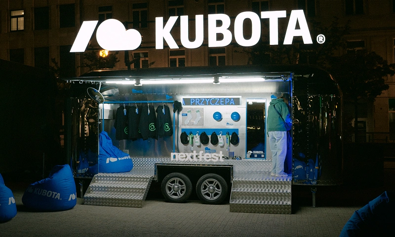 Baner podstrony Kubota na NEXT FEST. Mobilna przyczepa Kuboty, w której sprzedawane są festiwalowe produkty, takie jak ubrania i akcesoria.