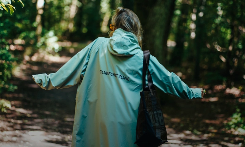 Baner kategorii moda wiosna lato 2026. Osoba widoczna od tyłu idzie leśną ścieżką w jasnozielonej parce przeciwdeszczowej z dużym napisem Comfort Zone na plecach i z ciemną torbą shopper na ramieniu.