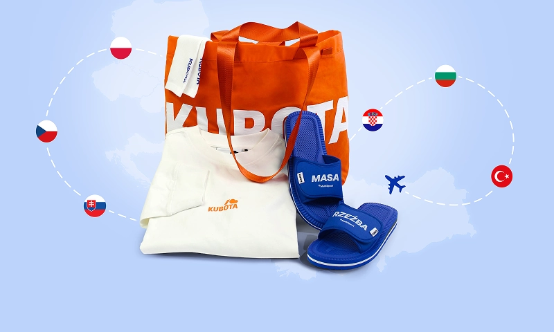 Baner podstrony Kubota x MultiSport. Sportowy zestaw podróżny składający się z pomarańczowej torby shopper, białej bluzki z długim rękawem oraz niebieskich klapków z motywacyjnymi napisami Masa i Rzeźba. Produkty są ułożone na jasnoniebieskim tle z motywem mapy i flagami państw połączonymi linią lotniczą.