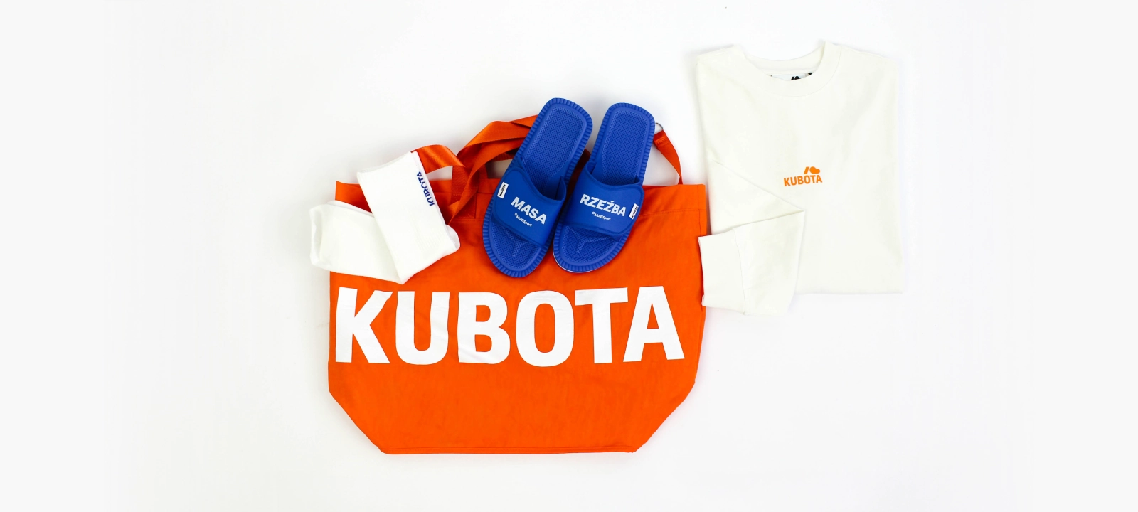 Zestaw kultowych gadżetów i odzieży lifestylowej ułożony w estetyczną kompozycję typu flat lay na czystym, białym tle. Głównym elementem zestawu jest pojemna, jaskrawo pomarańczowa torba shopper z dużym znakiem firmowym, na której umieszczono niebieskie klapki Rzep z motywacyjnymi hasłami Masa oraz Rzeźba. Obok torby leży biała bluzka z długim rękawem z drobnym logiem Kuboty oraz dopasowane sportowe skarpetki. Te wyjątkowe produkty promocyjne brały udział w wspólnej akcji konkursowej Kubota x MultiSport, która skutecznie promowała aktywny tryb wyjazdowy, zdrowe nawyki oraz sportowy styl życia.