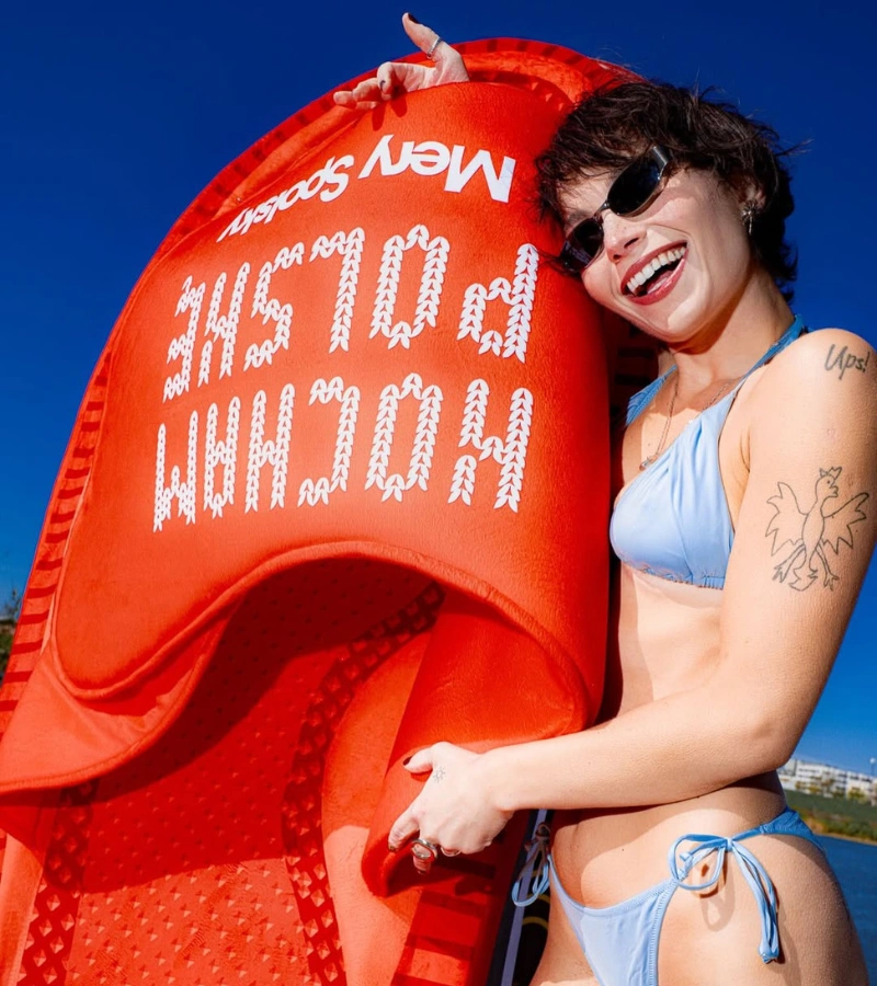 Zdjęcie @meryspolsky w jasnoniebieskim bikini i przeciwsłonecznych okularach obejmującej olbrzymi, limitowany klapek Kubota. Produkt w intensywnym czerwonym kolorze zdobiony jest białym, blokowym napisem Kocham Polskę oraz Mery Spolsky.