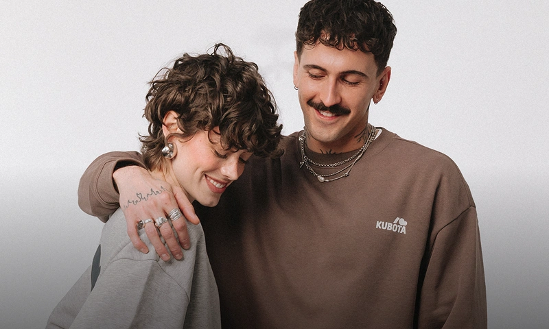 Uśmiechnięta, objęta para w modnych stylizacjach streetwear na jasnym tle. Mężczyzna ma na sobie brązową bluzę i wyraźnym napisem „Kubota”, a kobieta szarą wersję tej samej bluzy. Odkryj najchętniej kupowane modele klapków i ubrań.