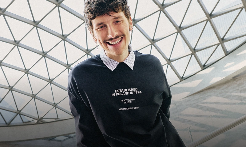 Uśmiechnięty mężczyzna pozuje na tle przeszklonej konstrukcji dachu w granatowym longsleeve z białymi nadrukami „Established in Poland in 1994" "Reactivated in 2018" i "Rebranded in 2025". Baner promuje kategorię nowości.